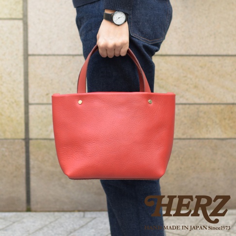 紅色職人手作皮革極簡托特包，配有兩個短提把和兩個金色铆釘，包身呈現細膩的皮革紋理，由穿著牛仔褲和深藍色毛衣的人士手持。