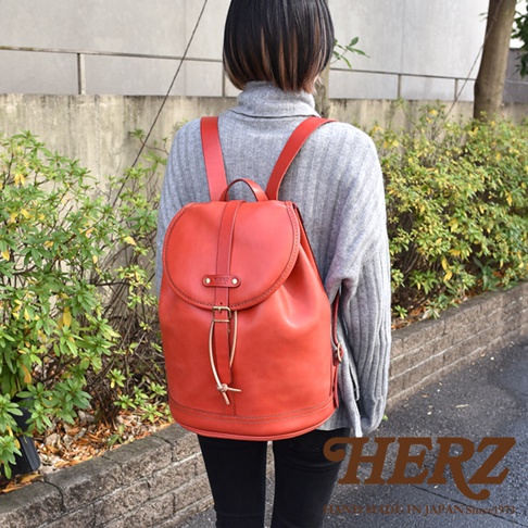 一位女性身穿灰色高領毛衣，背著一個紅色職人手作皮革單扣束口後背包。背包採用單扣設計，束口處有繩子和金屬扣環，皮革質樸有質感。