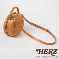 日本HERZ 職人手作皮革兩用圓形包，棕色皮革材質，附有可調整的肩背帶，細節處可見品牌標誌「HERZ」及「HAND MADE IN JAPAN Since1973」。