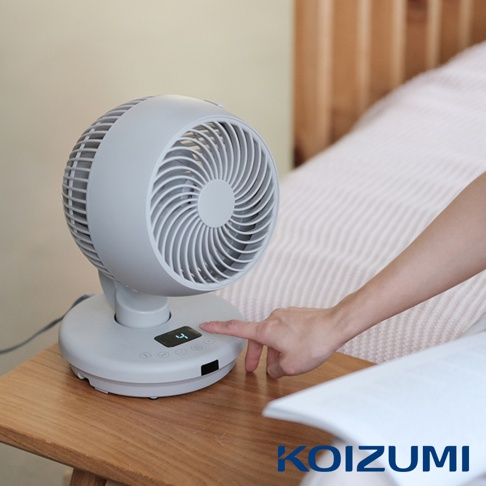 淺灰色鵝頸循環扇，帶有數字顯示螢幕，展示風速「4」，置於木桌上，旁邊是床鋪，一隻手正準備操作風扇。