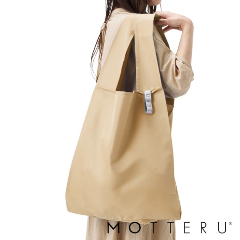 一位女性肩背一個淺卡其色的大容量摺疊環保隨行袋，袋子由尼龍材質製成，有兩個寬大的提把，袋身上有一個淺藍色的品牌標籤。