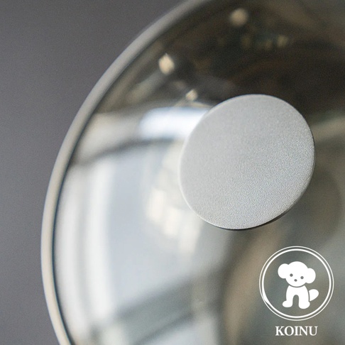 三層不鏽鋼雪平鍋的鍋蓋特寫，鍋蓋為金屬材質，帶有一個圓形銀灰色鍋鈕，鍋鈕表面有細緻顆粒感。右下角有白色小狗圖案的品牌標誌及「KOINU」字樣。