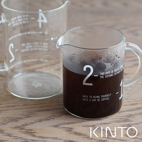 透明玻璃咖啡沖泡壺組，包含一個裝有深色咖啡的帶把手沖泡壺，壺身上印有白色數字「2」和「1」及英文標語，以及一個較大的空量杯，兩個沖泡壺均有白色刻度標記。