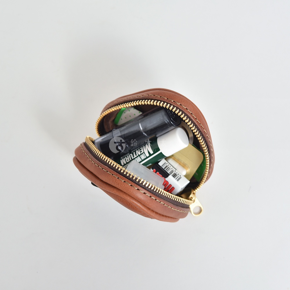 職人手作的棕色皮革三角飯糰造型小物收納包，拉鍊半開，內含各種小物，如藥膏、貼布等。