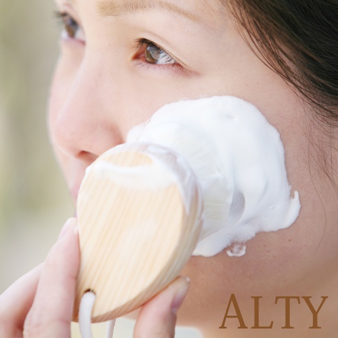 女性的臉頰上，一隻手握著木質的圓形洗臉刷，刷毛上沾滿了白色泡沫，正在清潔臉部。