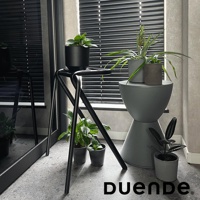 黑色金屬框架的吧檯椅，椅面上有盆栽植物，旁邊有一個灰色階梯狀的置物架，上面也有盆栽植物，背景是百葉窗和落地鏡。