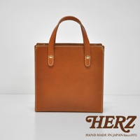 職人手作皮革直式托特包 T-44 XS，使用 tan 色澤的皮革製成，簡約的方形設計，配有兩條短提把，以白色縫線和金色金屬铆釘裝飾。