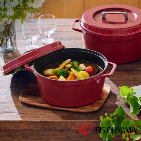 IRIS OHYAMA 萬用無水調理鍋，紅色鍋身內盛裝著燉煮蔬菜的料理，鍋蓋旁側擺放著酒杯及鮮花，背景為木質桌面。