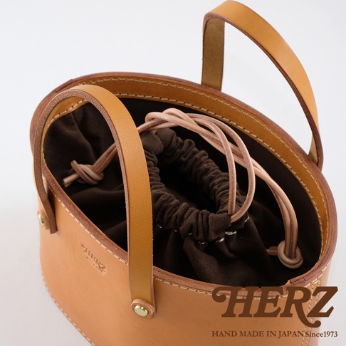 淺棕色皮革束口手提包，內襯為深棕色絨布，搭配同色系束繩。包身圓潤，提把為寬版皮革設計，整體風格簡約。
