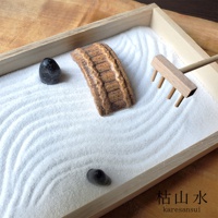 一個淺木框內，鋪滿白色細沙，沙面有規律的波紋，沙中擺放著一座褐色的石橋模型、兩顆黑色光滑的鵝卵石，以及一把木製的迷你耙子。