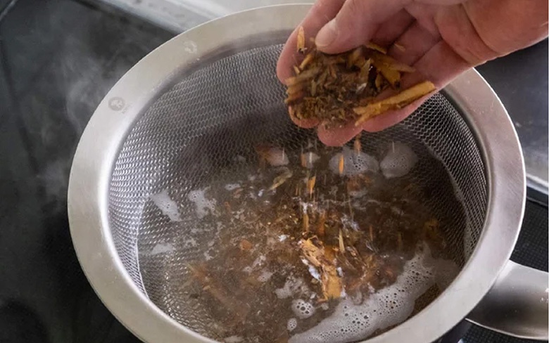 不鏽鋼濾水網與刻度料理盆，盆內裝有煮沸的水，一隻手正將乾燥的香草或食材倒入濾網中。