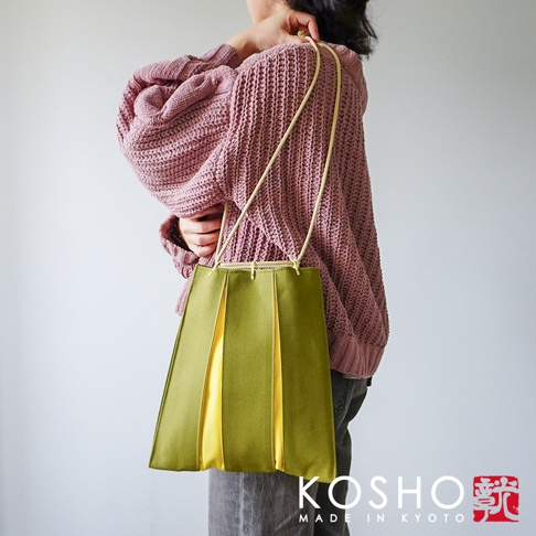 一位女性側身站立，背著一個來自京都KOSHO的ougi帆布信玄袋。該手袋以橄欖綠與亮黃色拼接呈現立體褶皺設計，搭配精緻的金色繩索提帶，整體風格簡約優雅，與模特身穿的淡紫色針織毛衣形成鮮明對比。