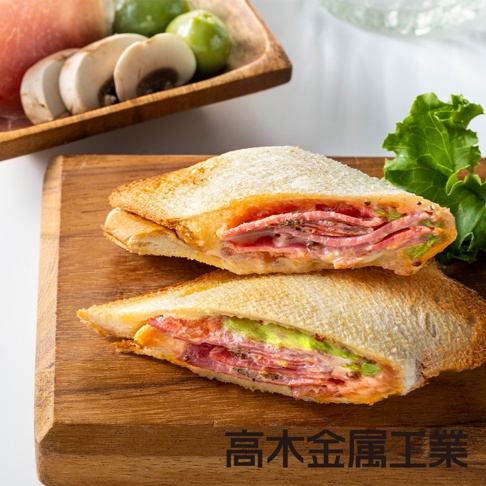 兩片烤過的口袋吐司，夾有火腿、番茄和醬料，擺放在木質砧板上，旁邊有生菜葉和一盤切片蘑菇、小番茄。