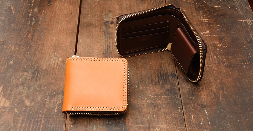 兩款日本HERZ職人手作皮革短夾置於木質桌面上，其中一個呈現淺棕色且拉鍊閉合，另一個則為深褐色並呈現開口狀態，展示內部的卡夾層與零錢袋設計。