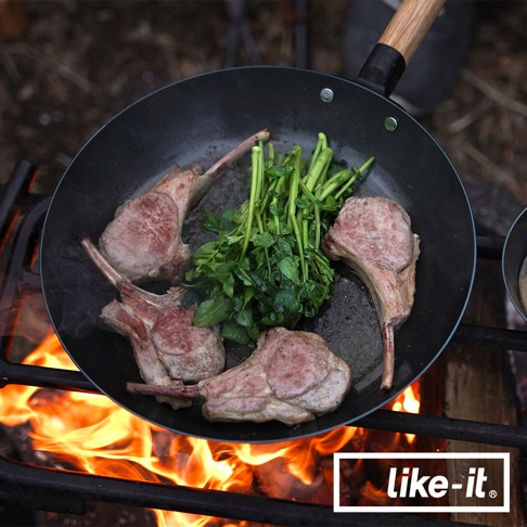 黑色鐵鍋中烹調著烤羊排與綠色蔬菜，鍋柄為木質，置於熊熊燃燒的營火上方。