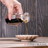 一隻手拿著裝有深色液體的透明玻璃醬油瓶，正將醬油倒入一個淺棕色的醬油碟中。瓶口有透明的塞子，醬油呈現琥珀色。背景為木紋。