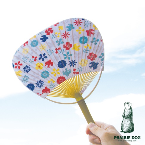 手持日式風情團扇，扇面為白色，印有紅、藍、黃、綠色的小花和鳥圖案，扇骨為黃色竹製，在藍天背景下展示。