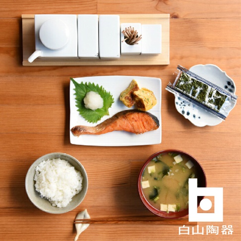 木質托盤上擺放著白色陶瓷調味罐、裝有烤鮭魚、玉子燒、蘿蔔泥和紫蘇葉的餐盤、盛有白飯的碗、味噌湯碗，以及一包海苔。