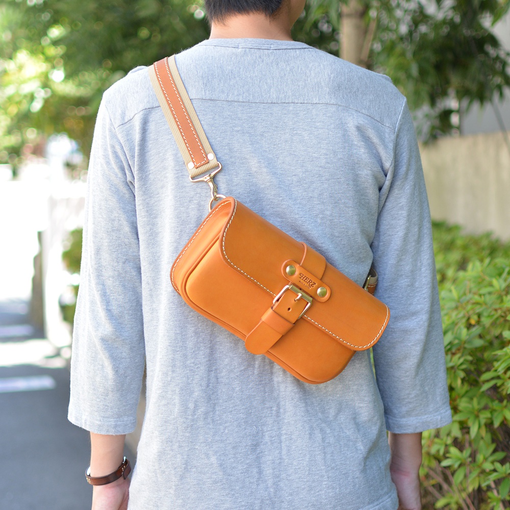 職人手作皮革單扣經典斜背包，呈現溫潤的橘棕色調，包身以光滑皮革製成，搭配米白色縫線。包款設有單一金屬扣環和皮質束帶，肩帶為織帶與皮革拼接設計，斜背於身。