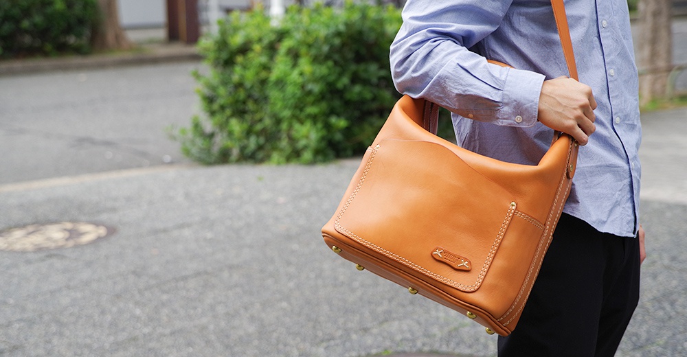 棕褐色皮革厚實單肩波士頓包，包身線條流暢，有明顯的縫線細節，包蓋上有品牌標誌，包底有四個金屬腳釘。