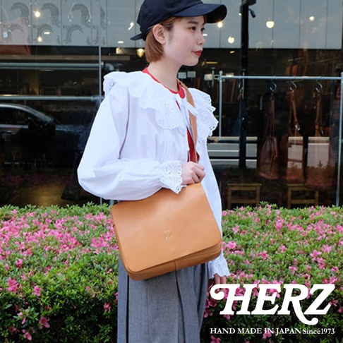 日本HERZ手工皮革品牌，一名女子身著白色荷葉領襯衫，搭配灰色裙子，肩背一個棕色軟皮斜背包，包上印有品牌標誌，背景是花叢和店鋪。