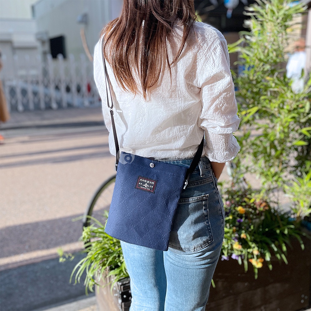 深藍色燈芯絨材質的平板收納側背包，搭配黑色細肩帶，正面有紅色標籤，由一位身穿白色蕾絲上衣和牛仔褲的女士斜背著。