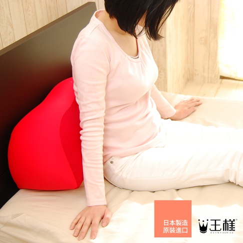 一個亮紅色的王樣膝下枕，呈梨形，放在臥室床鋪上，一位穿著粉色長袖上衣和白色褲子的女性坐在一旁。