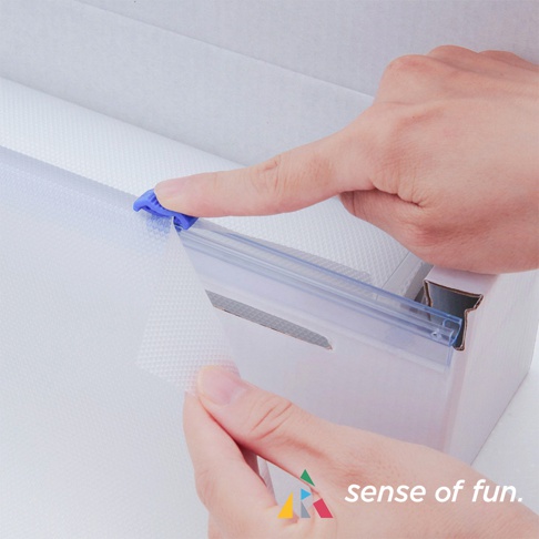 一隻手用藍色塑膠切刀切割白色帶紋理的捲筒式砧板保潔墊，墊片材質透明，長度約 800 公分。