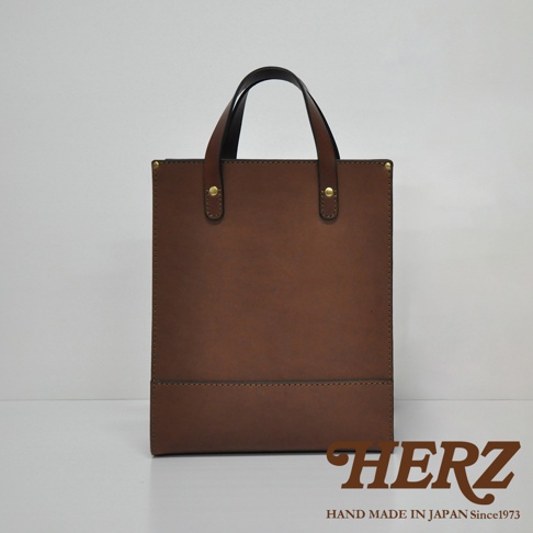 職人手作皮革直式托特包 T-44 M，採用溫潤的棕色皮革，簡約的方形設計，搭配一對皮質提把，提把處以金屬鉚釘固定，包身下方有一條橫向縫線裝飾，整體呈現經典實用的風格。