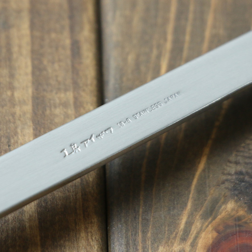一截銀色不鏽鋼萬用鍋鏟，上面刻有日文標誌和「18-8 STAINLESS JAPAN」字樣，背景為木紋桌面。