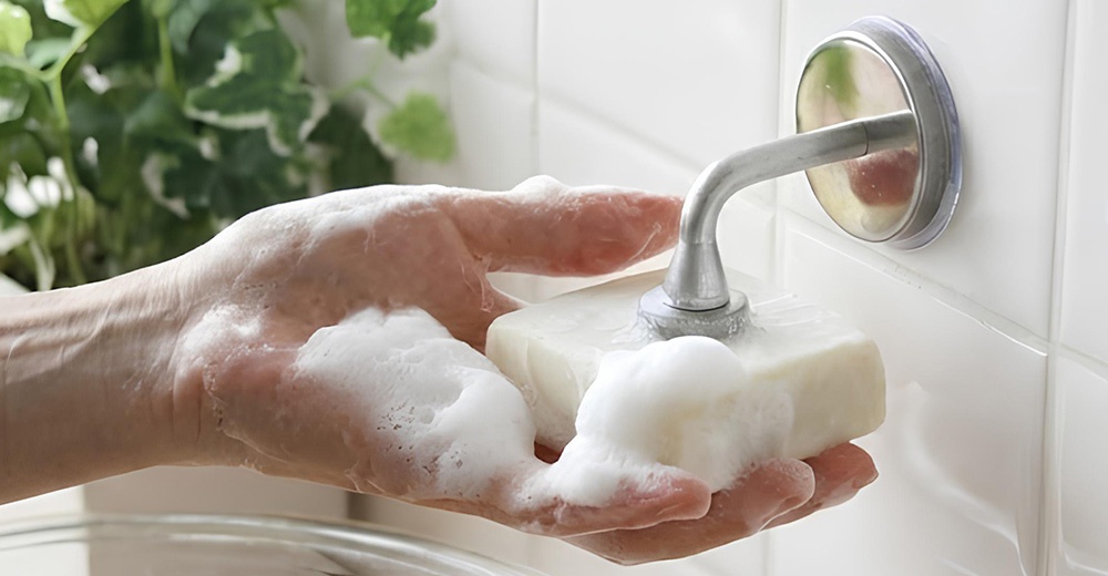 一個沾滿肥皂泡的手捧著一塊肥皂，旁邊是金屬製的肥皂架，背景是白色的磁磚牆和綠葉植物