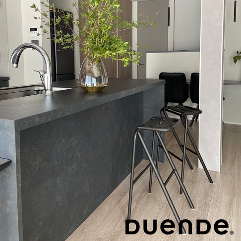 兩張黑色金屬框架的吧檯椅，椅面為黑色，椅腿呈細長交叉狀，擺放在深灰色石紋吧檯旁，地板為淺木紋。