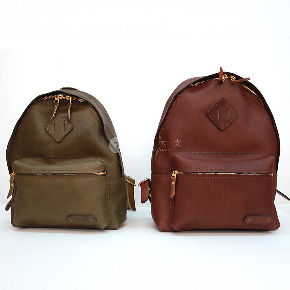 兩款職人手作皮革休閒風後背包，分別為橄欖綠和棕色。兩款背包均為圓潤的弧形設計，配備金色拉鍊、前置拉鍊口袋，並有品牌標誌。