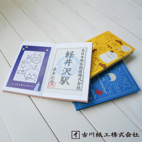 打開的御朱印帳，左頁有藍色背景的可愛卡通插圖，右頁是日文印章和文字。後方堆疊著一本黃色封面印有貓咪圖案和花朵的筆記本，以及一本藍色封面印有月亮、太陽和魚圖案的筆記本。