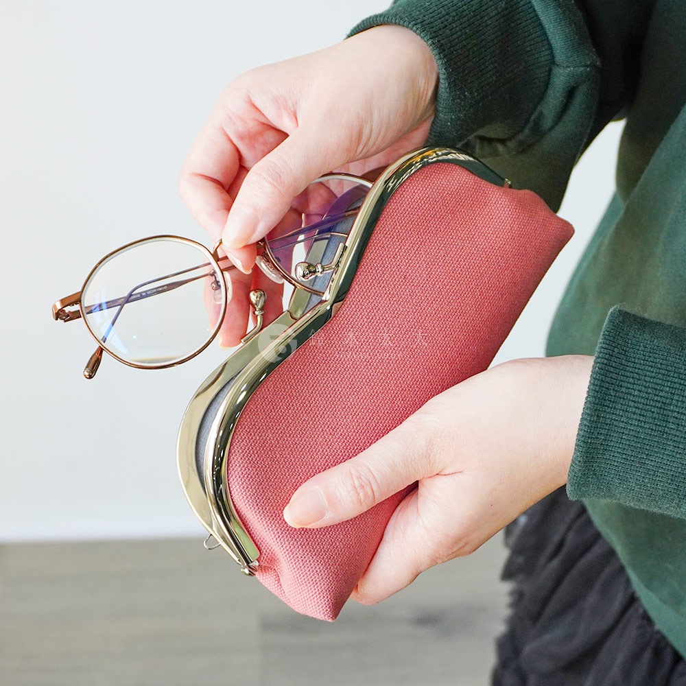 一個穿著綠色毛衣的人手裡拿著一個粉紅色口金眼鏡收納盒，並將一副金屬框眼鏡放入其中。