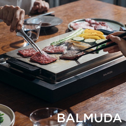 黑色極致電烤盤，烤盤上擺放著多塊生牛肉、烤過的肉片、玉米筍、櫛瓜和香菇。左手持金屬夾子，右手持黑色筷子，正在翻動烤盤上的食材。