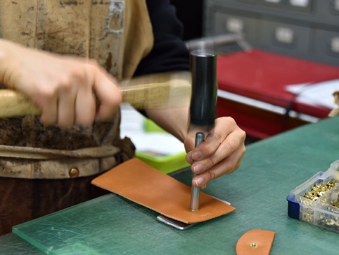 雙折皮革零錢包製作過程中，師傅正使用金屬壓具和木槌在淺棕色皮革上進行壓孔作業，一旁有裝有鉚釘的收納盒。