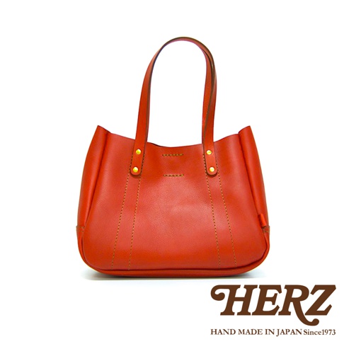 橘紅色皮革手提包，附雙提把，包身線條簡潔，綴有金色鉚釘與縫線細節，底部圓潤。