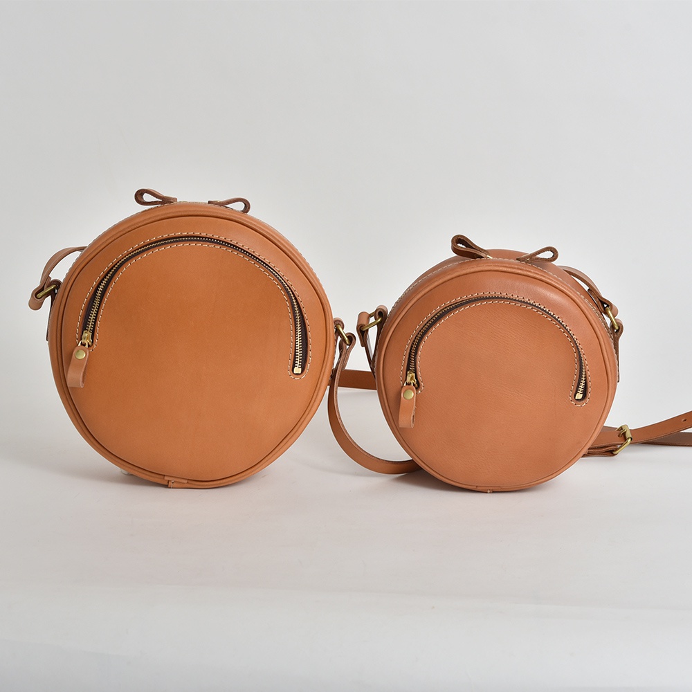 職人手作皮革小圓包 CK-19 M，以天然皮革製成，呈現溫潤的棕色調。圓潤的包身設計，搭配弧形拉鍊開口，簡約大方。包款頂部裝飾有皮革蝴蝶結，增添精緻細節。