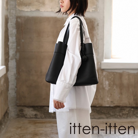 一位女性側面站立，肩背著一只極簡風格的 itten-itten 黑色義大利皮革托特包，包身表面具有細膩的壓紋質感，側邊飾有精緻的皮革繩結吊飾，她身穿寬鬆的純白長袖襯衫與長褲，背景為帶有復古質感的灰褐色牆面。