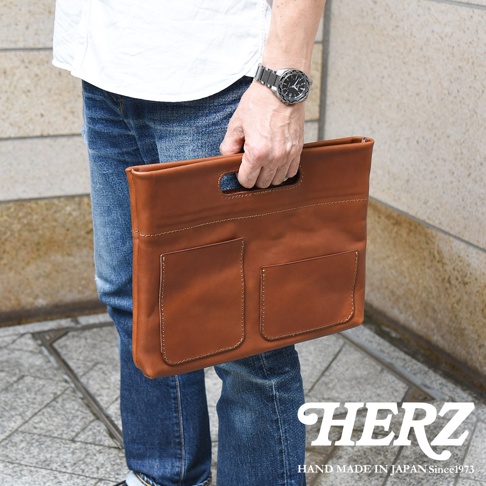 一位穿著白色上衣與藍色牛仔褲的男士，手持一個由日本HERZ品牌職人手作的棕色皮革手拿包，包款正面設有兩個外袋與簡約的提把設計，整體呈現出經典且實用的皮革質感。