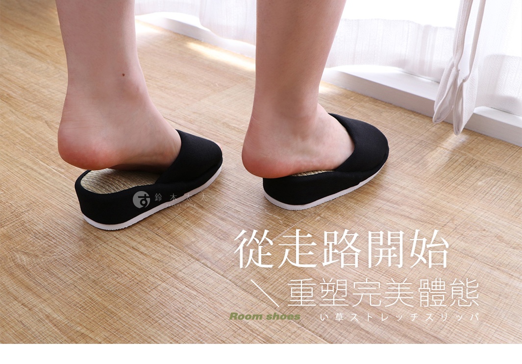 一雙黑色藺草材質的室內拖鞋，鞋面為織物材質，鞋底為白色。一雙腳穿著拖鞋，腳後跟抬起，露出部分藺草內墊。