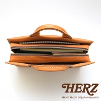 日本HERZ手工皮革簡約公事包，頂部視角，皮革顏色為棕色，包內裝有文件夾和筆記本，並有品牌HERZ和HAND MADE IN JAPAN Since1973的標誌。