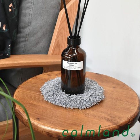 圓形灰色絨毛植物盆栽墊，質感柔軟，尺寸適合中型盆栽，置於木質圓桌上，周圍有擴香瓶與木質椅凳。