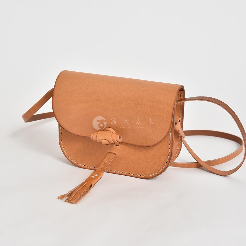 職人手作皮革流蘇側背包，呈現天然棕色皮革，包身帶有圓潤弧線，並有精緻的流蘇與繩結飾扣，簡約而有質感。