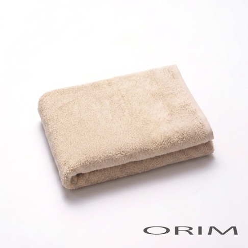 一張對摺擺放的淺米色 ORIM 品牌 PLUME 系列浴巾，呈現柔軟蓬鬆的純棉質感，右下方印有深色的 ORIM 品牌標誌，整體背景為乾淨純白。