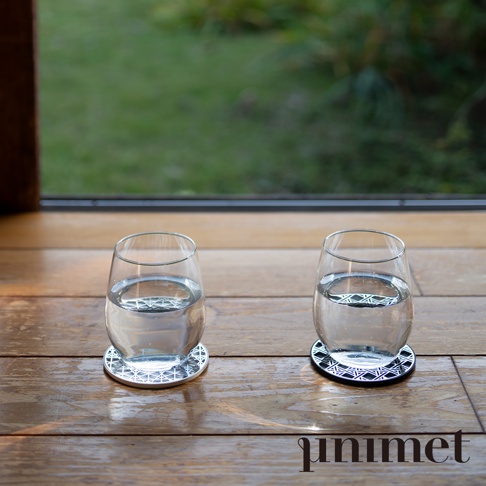 木質地板上擺放著兩個裝有清水的透明玻璃杯，杯底各墊著一個 unimet 不鏽鋼光影藝術防滑杯墊，左側為銀色款，右側為黑色款，杯墊表面呈現精緻的幾何鏤空圖案，背景為模糊的戶外綠色庭院景緻。