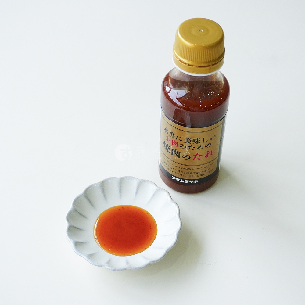 一瓶金蓋封口的朝紫燒肉沾醬放置在純白背景上，旁邊的小巧白色花瓣造型碟子中盛裝著少許深琥珀色的醬汁，呈現出清晰的產品與醬料色澤。