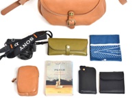 一個由棕色皮革製成的馬鞍包，搭配一個黑色索尼相機，一個橄欖綠色皮夾，一本日文書，一個棕色皮革手機包，一個黑色鑰匙包，一個黑色錢包，以及一個藍色印花手帕。