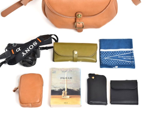 一個由棕色皮革製成的馬鞍包，搭配一個黑色索尼相機，一個橄欖綠色皮夾，一本日文書，一個棕色皮革手機包，一個黑色鑰匙包，一個黑色錢包，以及一個藍色印花手帕。